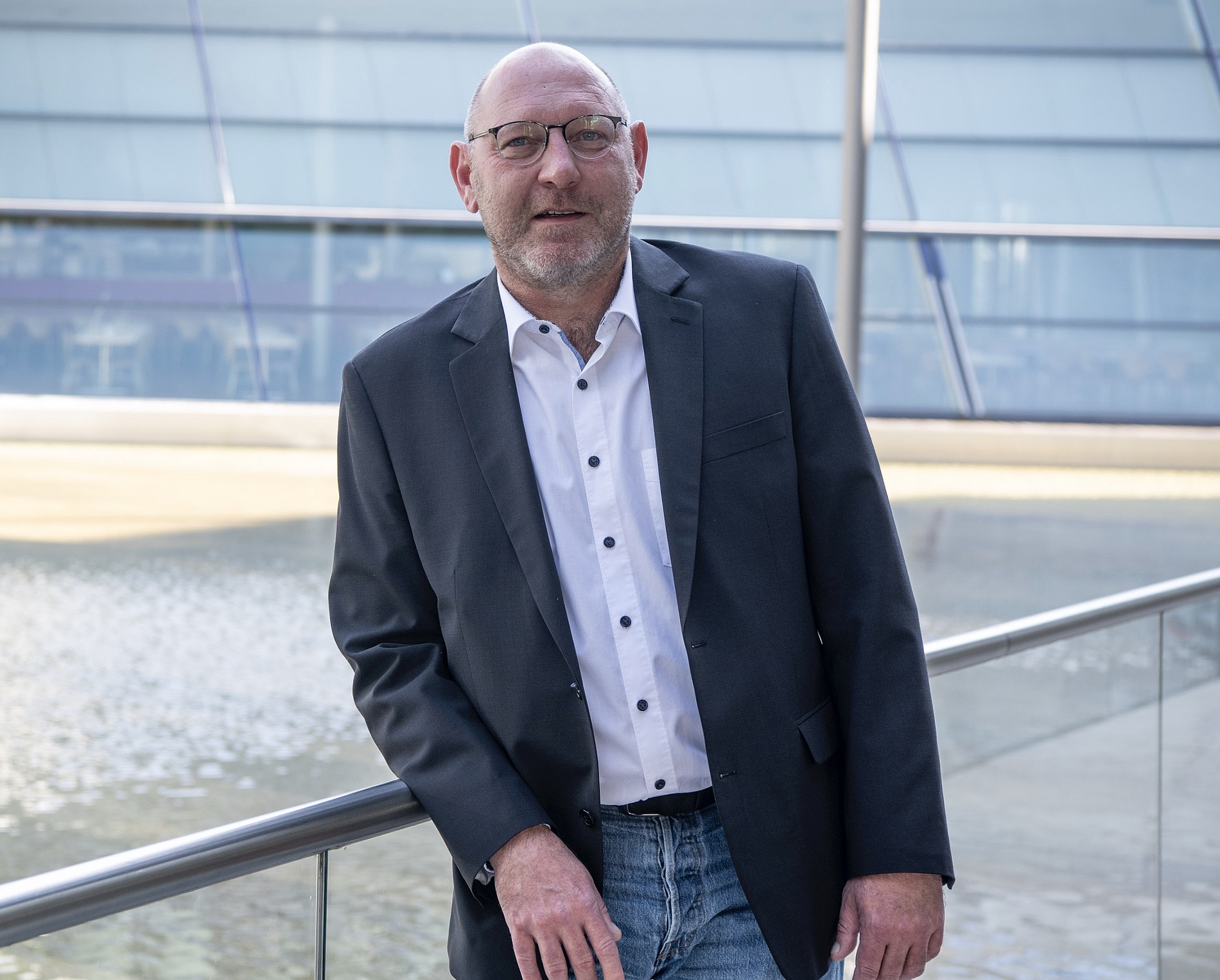 Kai Michael Dietrich, der eine Brille, ein weißes Hemd und einen dunklen Blazer trägt, steht an einem Geländer vor einem modernen Glasgebäude mit Wasserspiel.