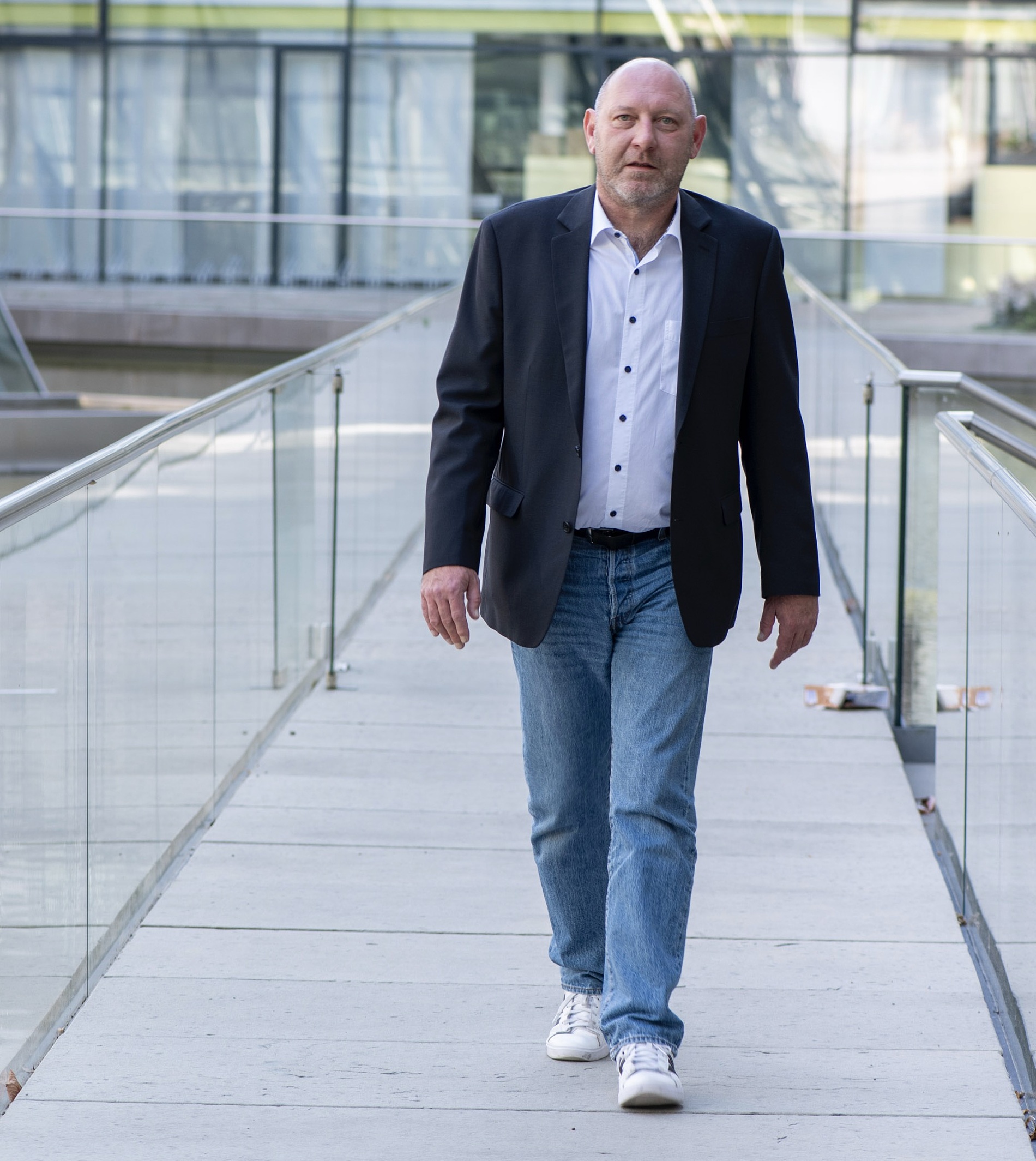Kai Michael Dietrich in Blazer, Jeans und Turnschuhen geht allein über eine moderne gläserne Fußgängerbrücke.