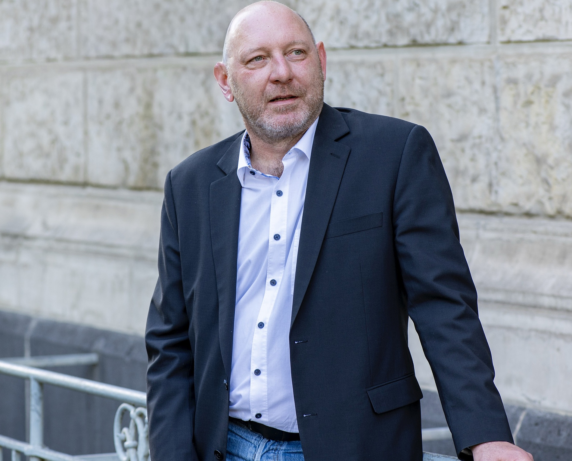 Kai Michael Dietrichmit schwarzem Blazer, weißem Hemd und Jeans steht im Freien vor einer Steinmauer.