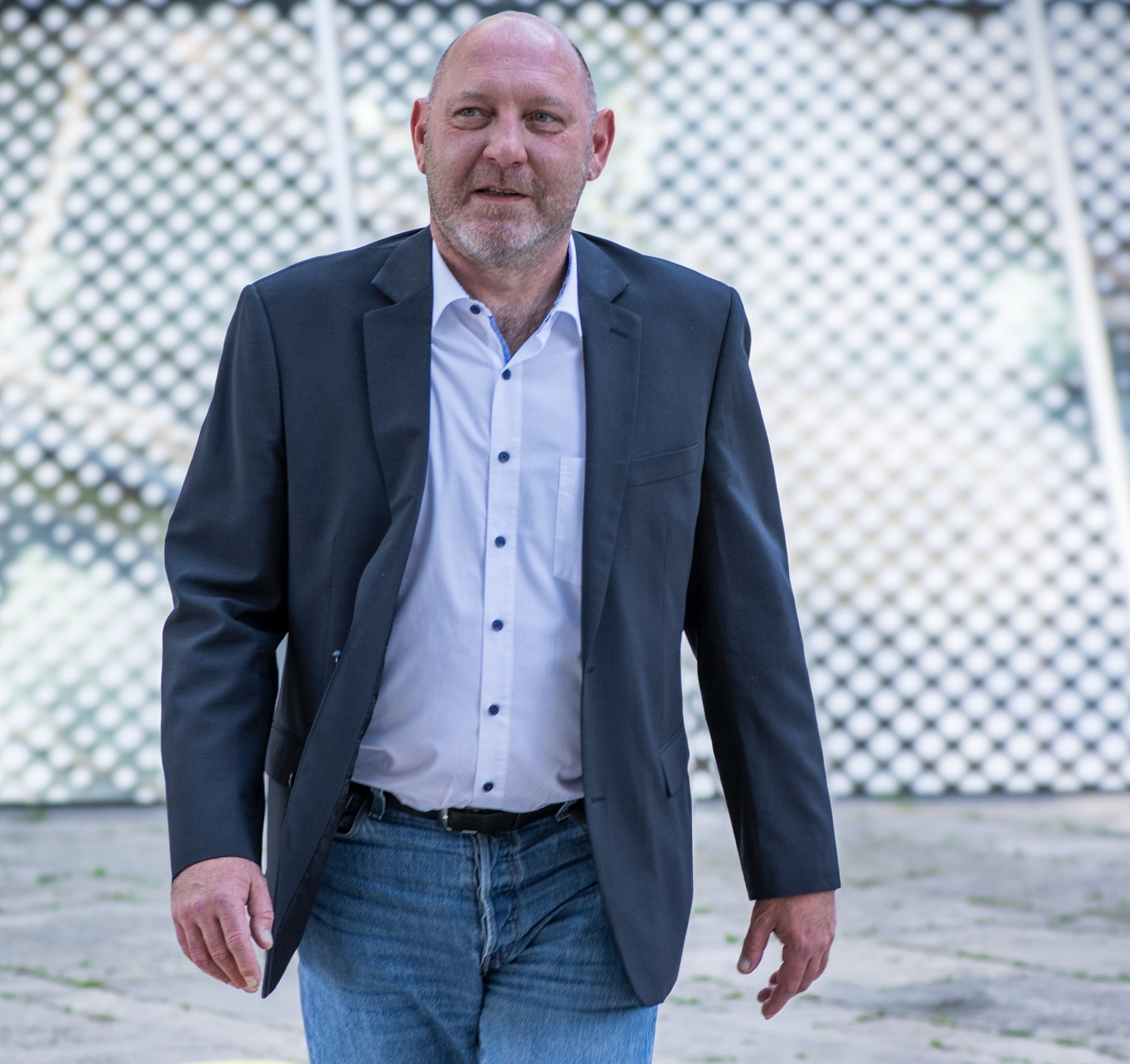 Kai Michael Dietrich in einem dunklen Blazer, weißem Hemd und blauen Jeans geht im Freien vor einem gemusterten Hintergrund spazieren.
