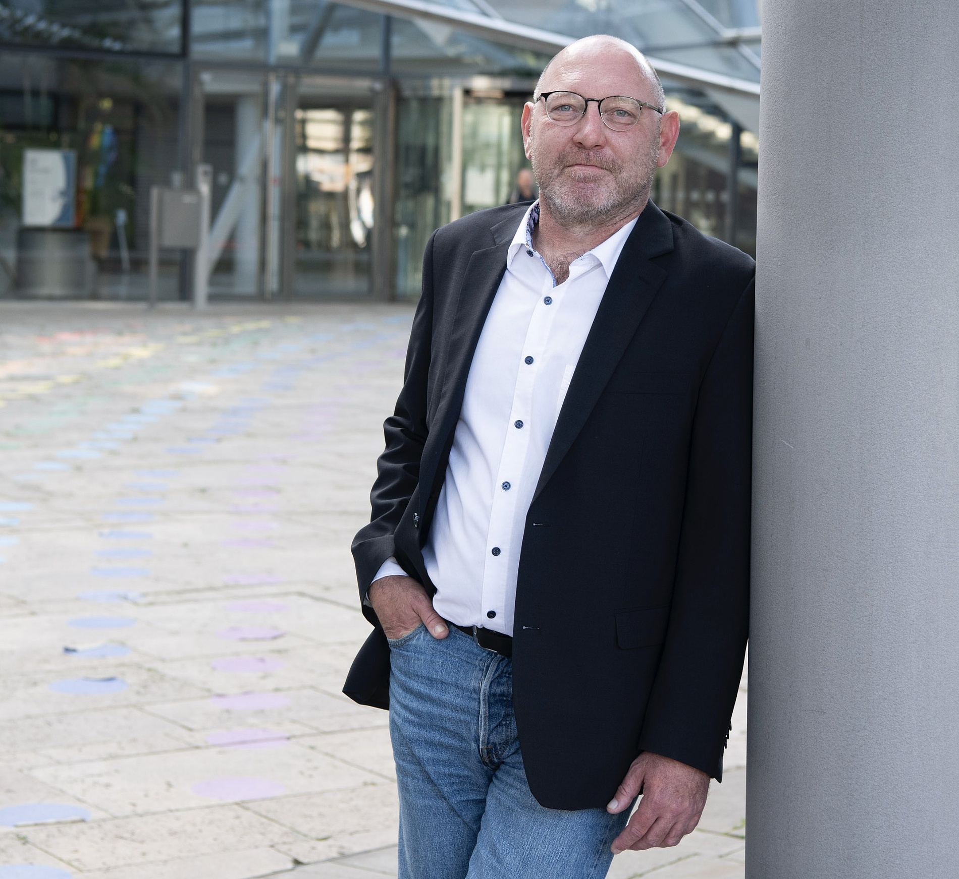 Kai Michael Dietrich mit Brille und Bart, der einen schwarzen Blazer, ein weißes Hemd und Jeans trägt, steht im Freien und lehnt sich an eine große Säule neben einem modernen Glasgebäude.