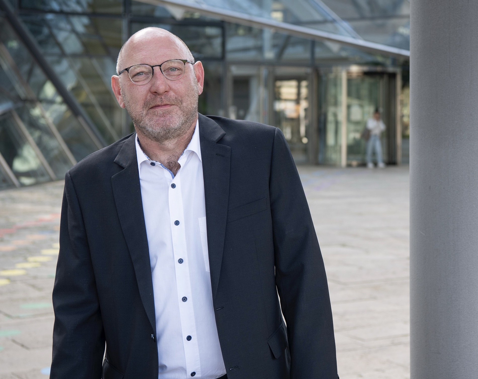 Kai Michael Dietrich im Anzug und mit Brille steht im Freien vor einem modernen Glasgebäude.