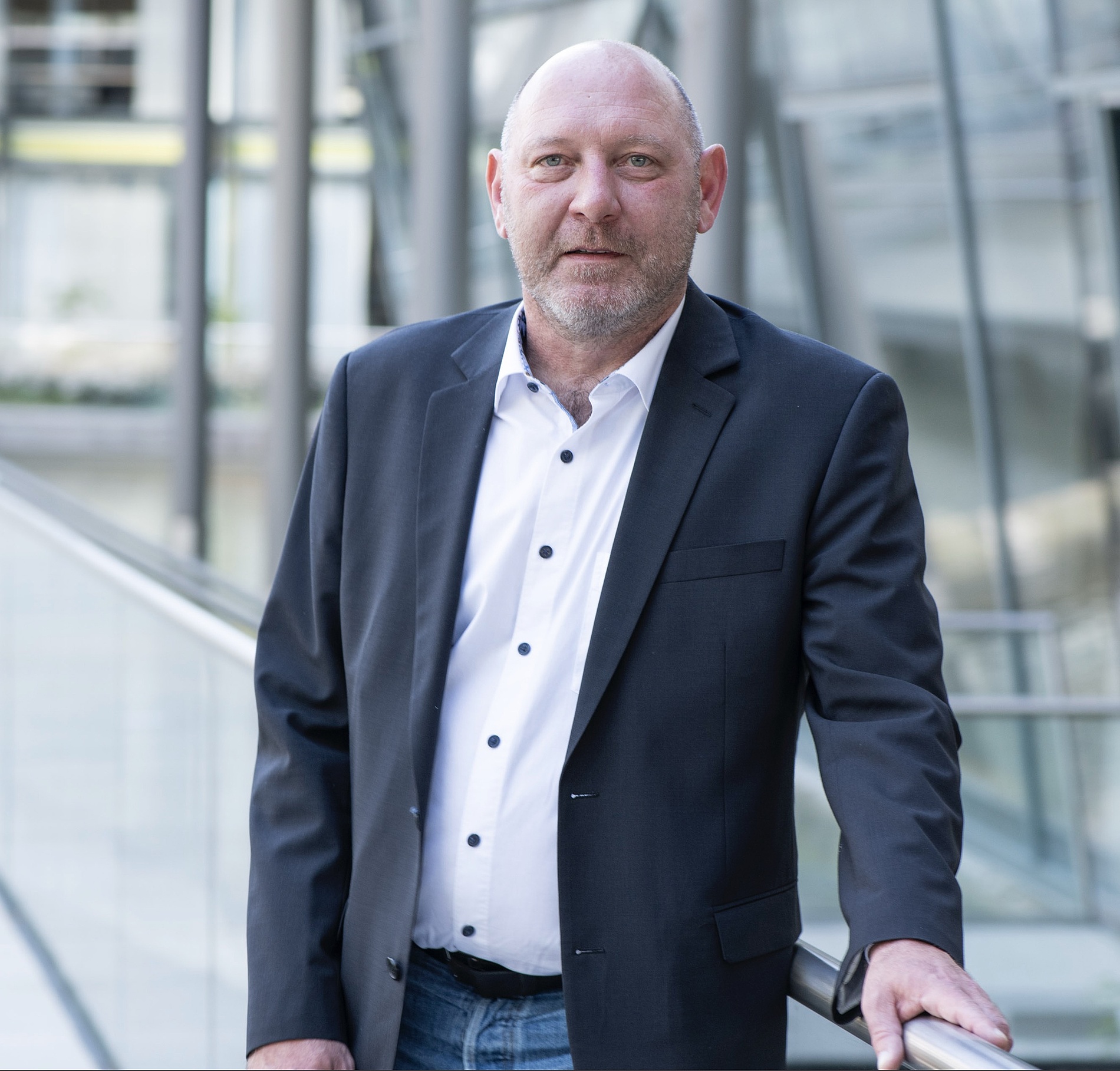 Kai Michael Dietrich mit dunklem Blazer und weißem Hemd steht in einem Haus und stützt sich auf ein Glasgeländer mit einem modernen Gebäude im Hintergrund.