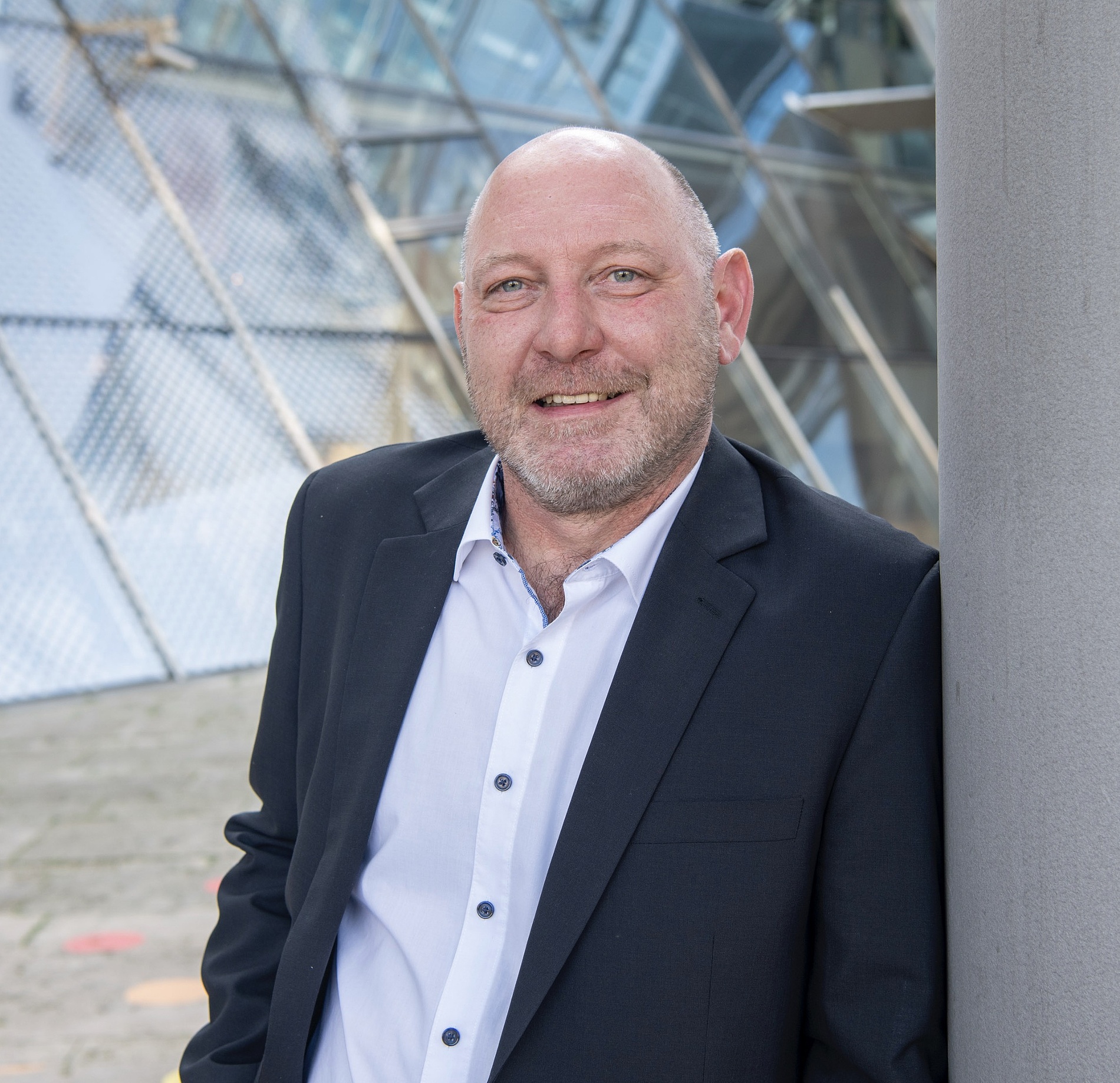 Kai Michael Dietrich  in Anzugjacke und weißem Hemd lehnt sich an eine Säule im Freien, mit moderner Glasarchitektur im Hintergrund.