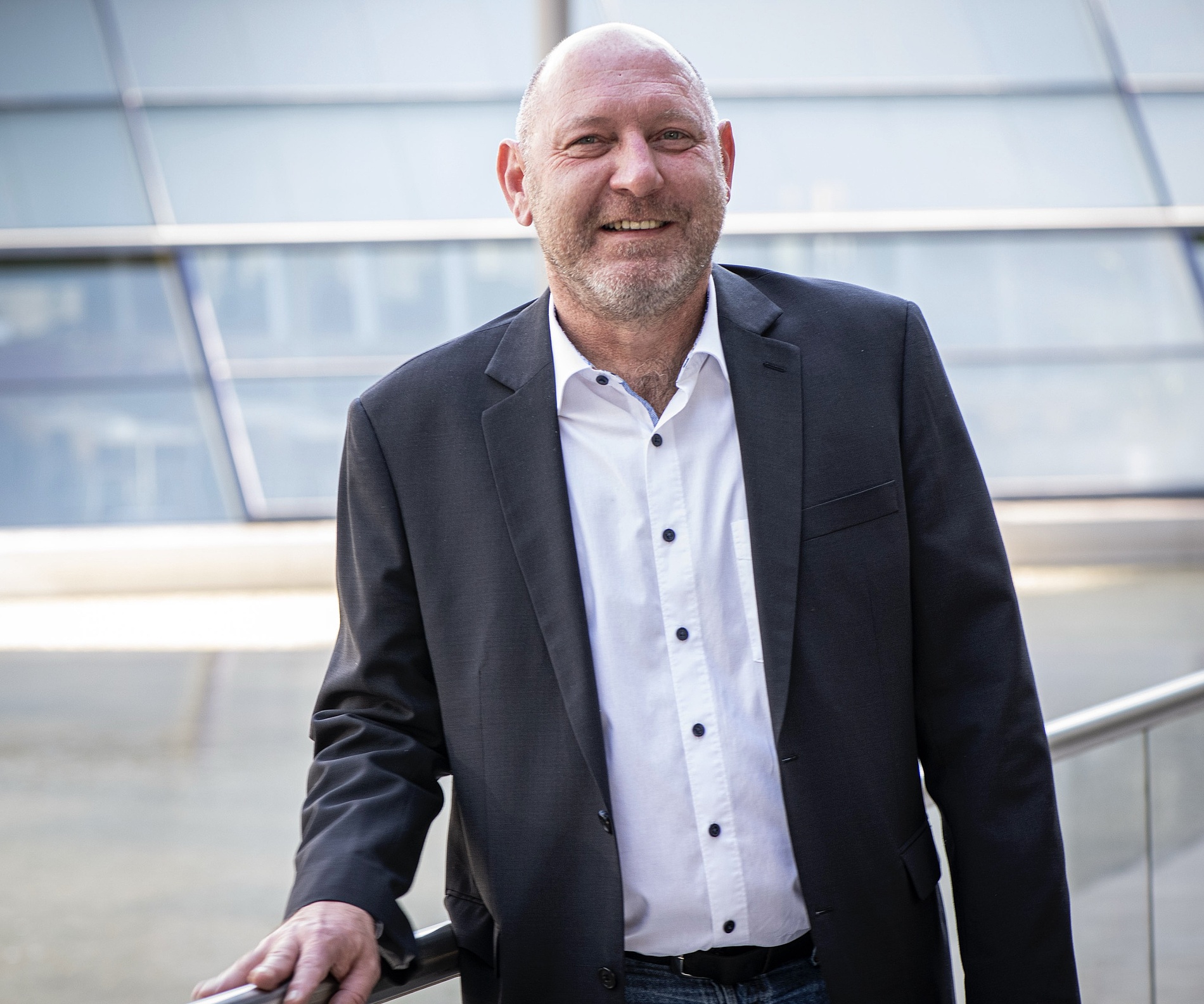 Kai Michael Dietrich in einem dunklen Blazer und einem weißen Hemd steht an einem Metallgeländer vor einem modernen Glasgebäude und lächelt in die Kamera.