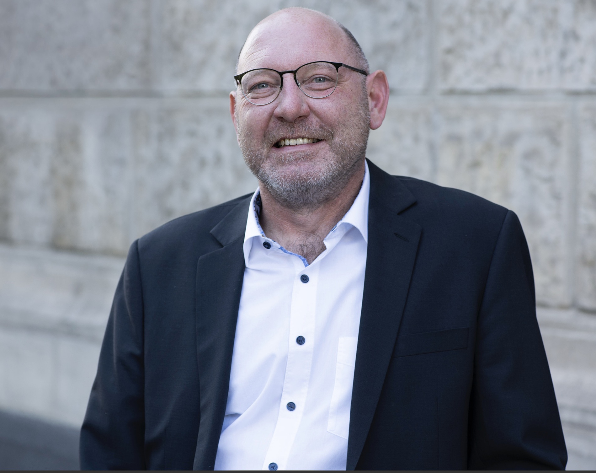 Kai Michael Dietrich schwarzem Anzug und weißem Hemd, steht vor einer strukturierten Steinmauer und lächelt in die Kamera.