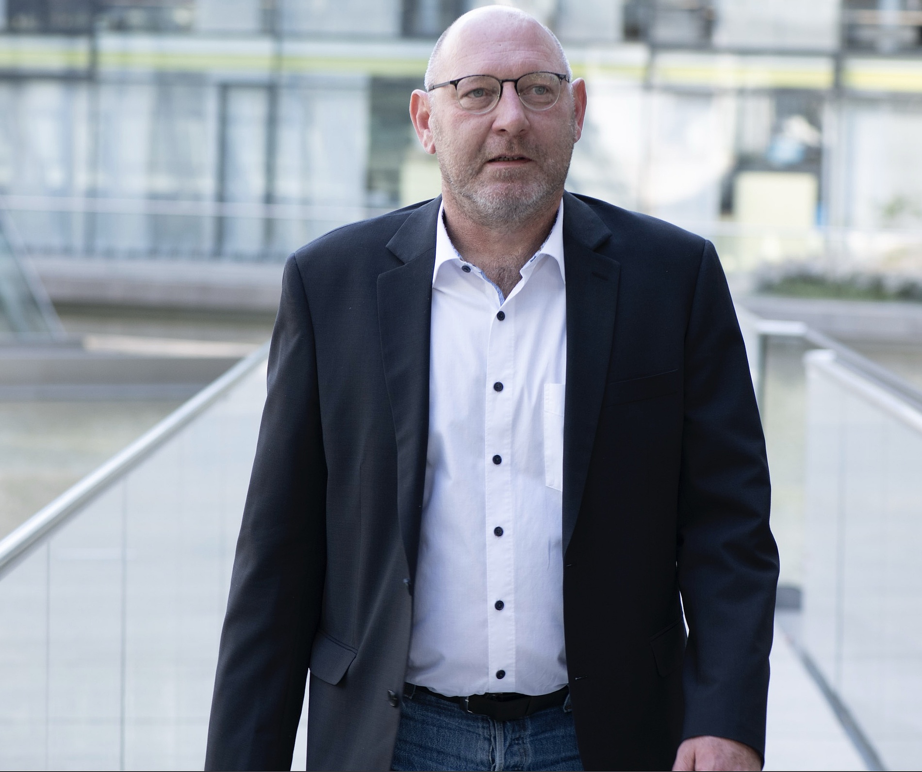 Kai Michael Dietrich, weißem Hemd und dunklem Blazer geht im Freien auf einer Fußgängerbrücke mit modernen Gebäuden im Hintergrund.