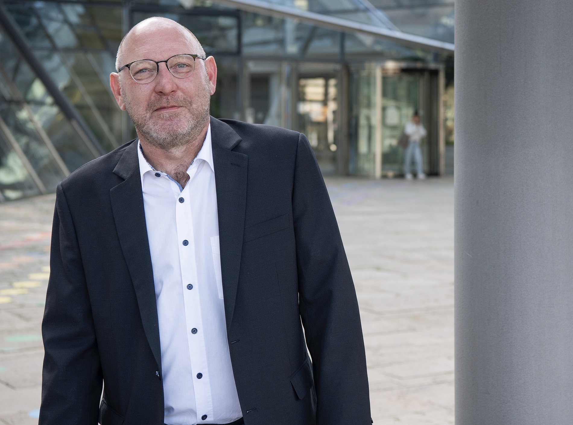Kai Michael Dietrich im Anzug und mit Brille steht im Freien neben einem modernen Glasgebäude und blickt in die Kamera.