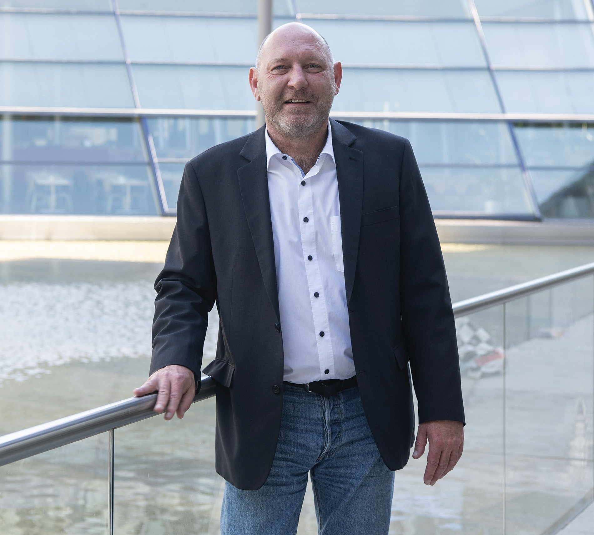 Kai Michael Dietrich mit weißem Hemd und Jeans steht an einem Geländer vor einem modernen Glasgebäude mit Wasserspiel.