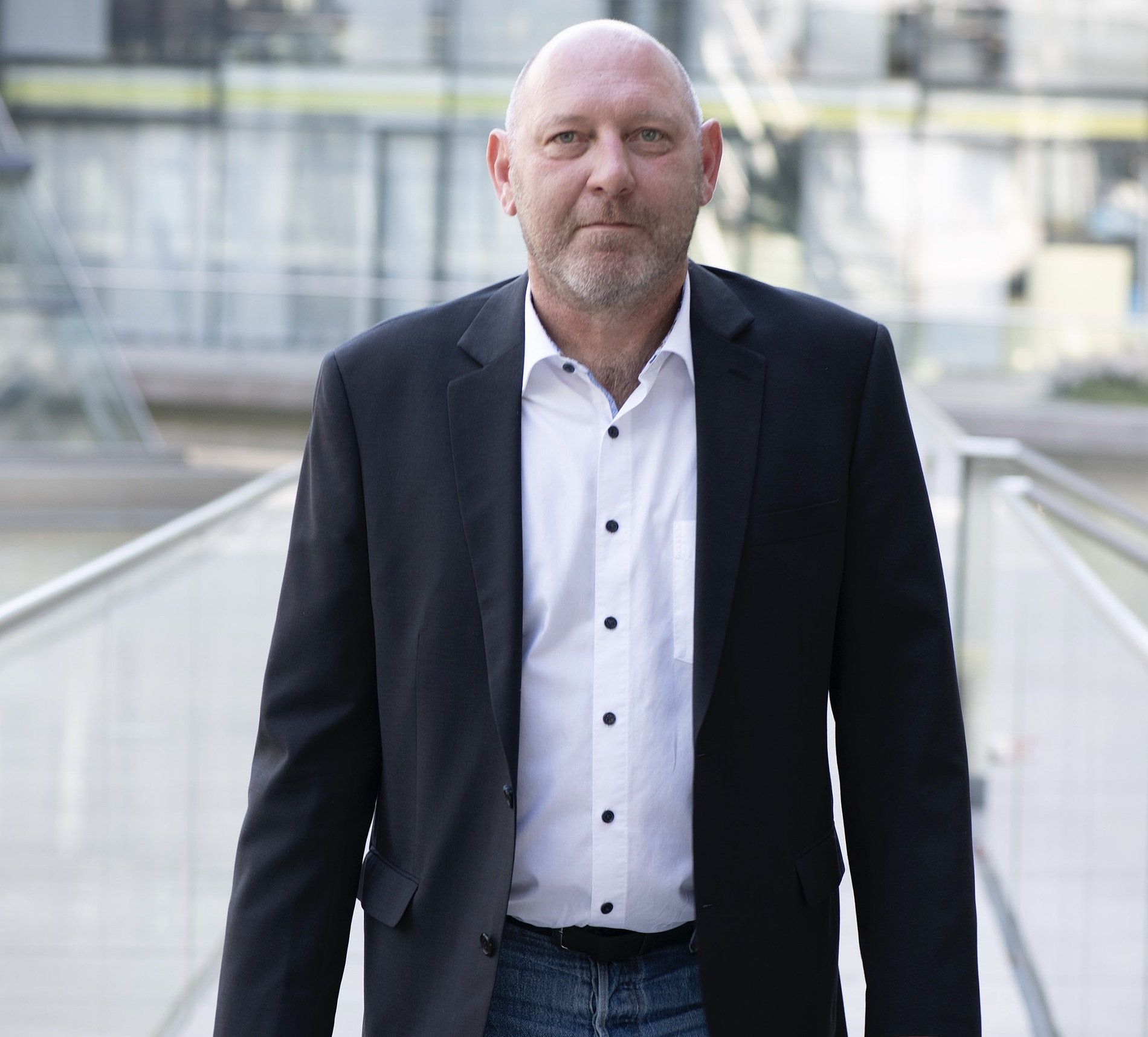 Kai Michael Dietrich und gestutztem Bart, der einen schwarzen Blazer, ein weißes Hemd und Jeans trägt, steht im Freien auf einer Fußgängerbrücke.