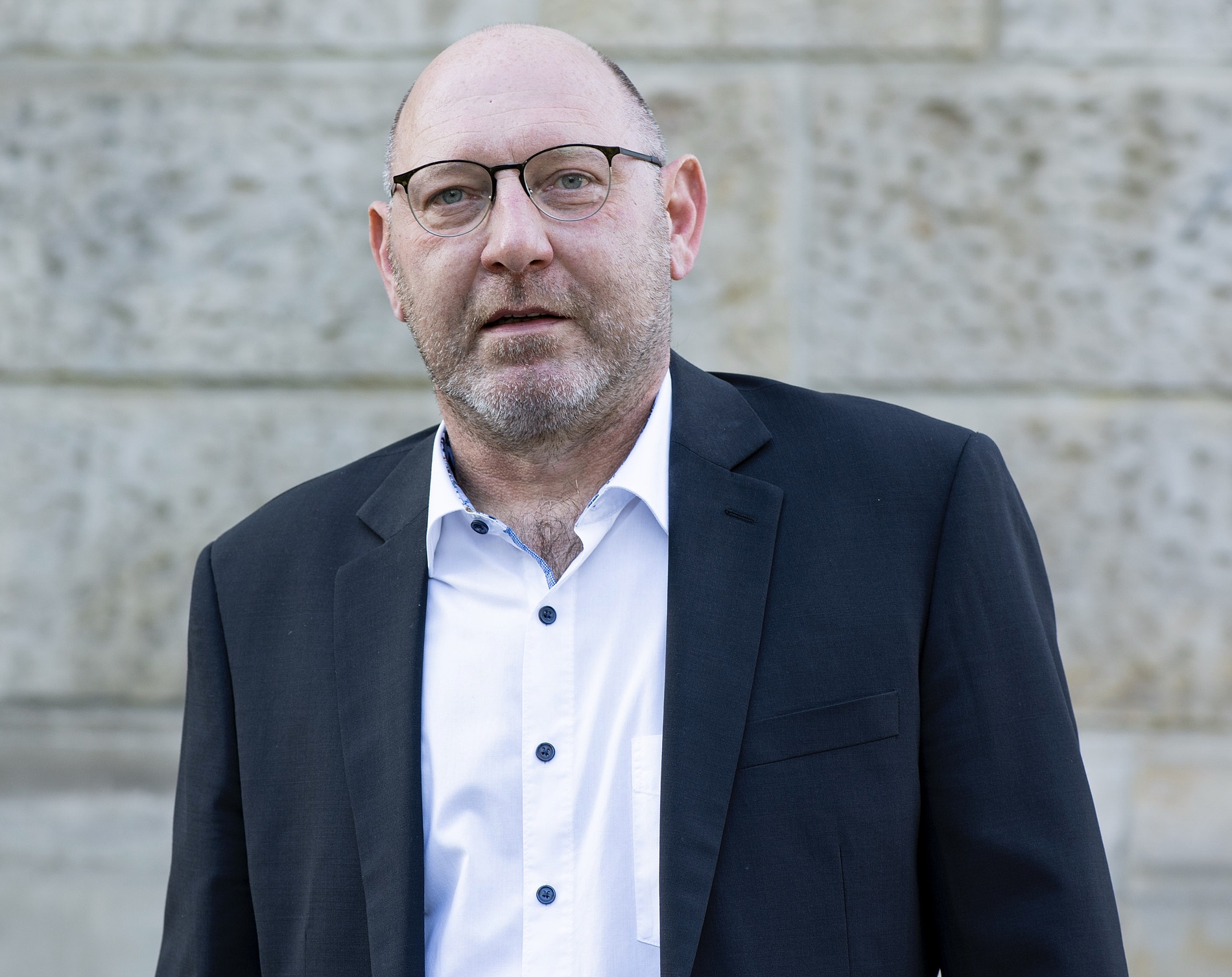 Kai Michael Dietrich, der eine Brille, ein weißes Hemd und einen dunklen Blazer trägt, steht vor einer Steinmauer.