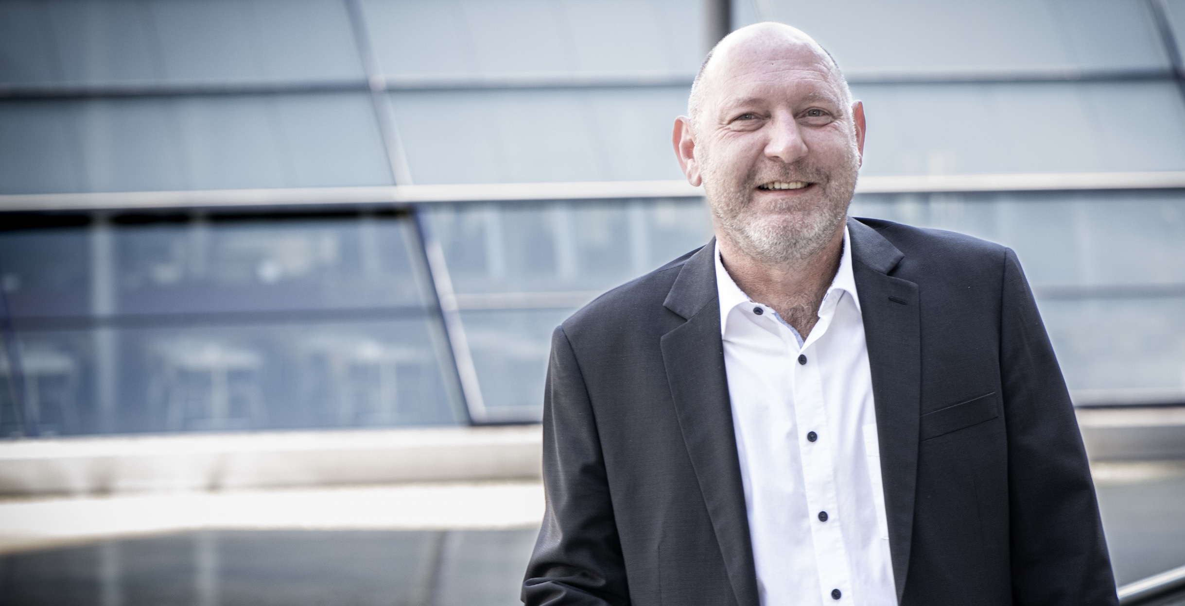 Rechtsanwalt Kai Michael Dietrich im Anzug steht im Freien vor einem modernen Glasgebäude und lächelt in die Kamera.